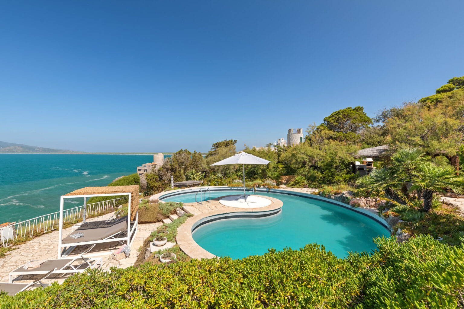 Scenic view of a luxury villa poolside overlooking the turquoise ocean, featuring a circular swimming pool, sun loungers, and a shaded cabana, surrounded by lush greenery and clear blue skies. Perfect for relaxing and enjoying coastal views. | Only on SopranoVillas