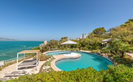 Scenic view of a luxury villa poolside overlooking the turquoise ocean, featuring a circular swimming pool, sun loungers, and a shaded cabana, surrounded by lush greenery and clear blue skies. Perfect for relaxing and enjoying coastal views.
