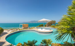 Scenic ocean view from a luxury villa with a circular swimming pool, lounge chairs, and a shaded area, surrounded by lush tropical plants and clear blue skies. Perfect for relaxation and enjoying coastal views.