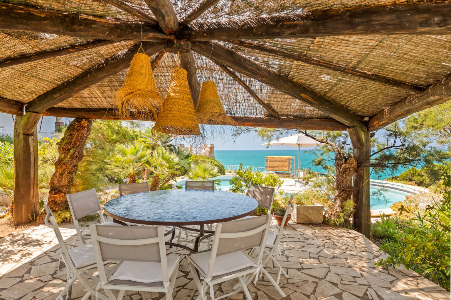 Outdoor dining area under a thatched roof, featuring a round table and white chairs, with a scenic view of a pool and the ocean in the background. Ideal for relaxing meals in a tranquil setting. | Only on SopranoVillas