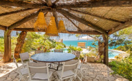 Outdoor dining area under a thatched roof, featuring a round table and white chairs, with a scenic view of a pool and the ocean in the background. Ideal for relaxing meals in a tranquil setting.