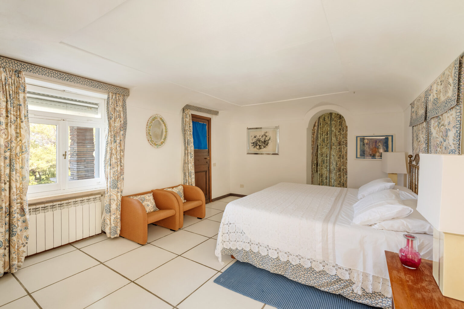 Cozy bedroom featuring a double bed, two armchairs, and natural light from a window. Decorated with floral curtains and artwork, the room offers a warm and inviting atmosphere. | Only on SopranoVillas