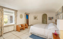 Cozy bedroom featuring a double bed, two armchairs, and natural light from a window. Decorated with floral curtains and artwork, the room offers a warm and inviting atmosphere.