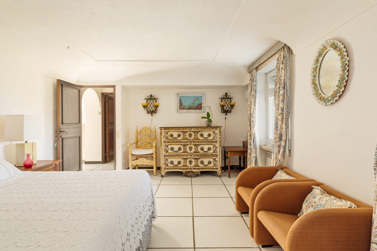 Cozy bedroom interior featuring a double bed, vintage dresser, and comfortable seating area with two armchairs. Natural light floods the space through an open door, highlighting the decor and tiled flooring. | Only on SopranoVillas
