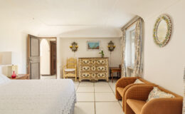 Cozy bedroom interior featuring a double bed, vintage dresser, and comfortable seating area with two armchairs. Natural light floods the space through an open door, highlighting the decor and tiled flooring.