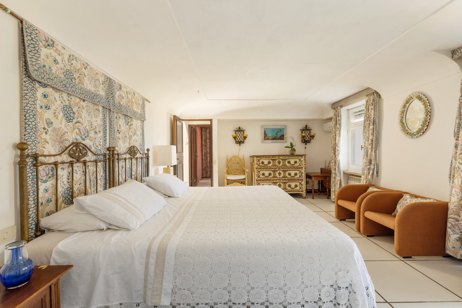 Cozy bedroom featuring a large bed with a white lace cover, elegant wall decor, and vintage furniture, including a dresser and seating area, creating a warm and inviting atmosphere. | Only on SopranoVillas
