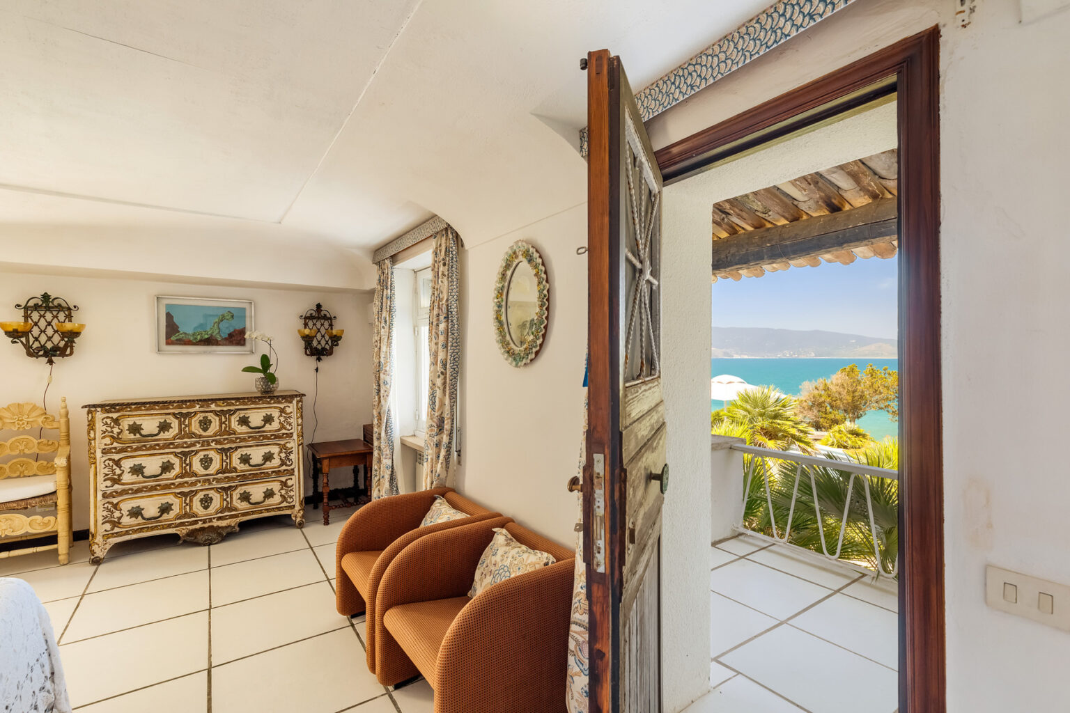Cozy interior of a seaside vacation rental featuring a view of the ocean from an open door, with stylish furniture including two armchairs and a decorative dresser. | Only on SopranoVillas
