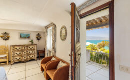 Cozy interior of a seaside vacation rental featuring a view of the ocean from an open door, with stylish furniture including two armchairs and a decorative dresser.