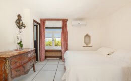Cozy bedroom featuring a white bed, vintage dresser, and large window with scenic views, complemented by soft curtains and air conditioning for comfort.