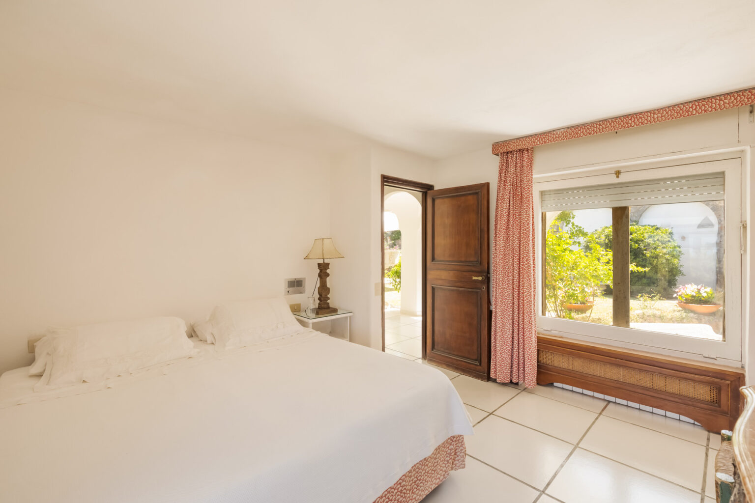 Bright and airy bedroom featuring a white bed, wooden furniture, and large windows with views of a garden. Natural light floods the space, highlighting the neutral color palette and simple decor. | Only on SopranoVillas