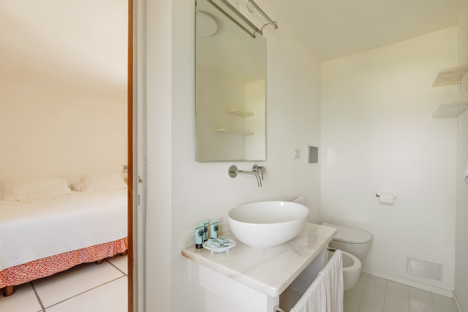 Modern bathroom with a round sink, mirror, and toilet, featuring neutral walls and a glimpse of an adjacent bedroom with a white bed and decorative bedspread. | Only on SopranoVillas