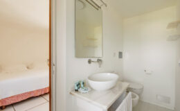 Modern bathroom with a round sink, mirror, and toilet, featuring neutral walls and a glimpse of an adjacent bedroom with a white bed and decorative bedspread.