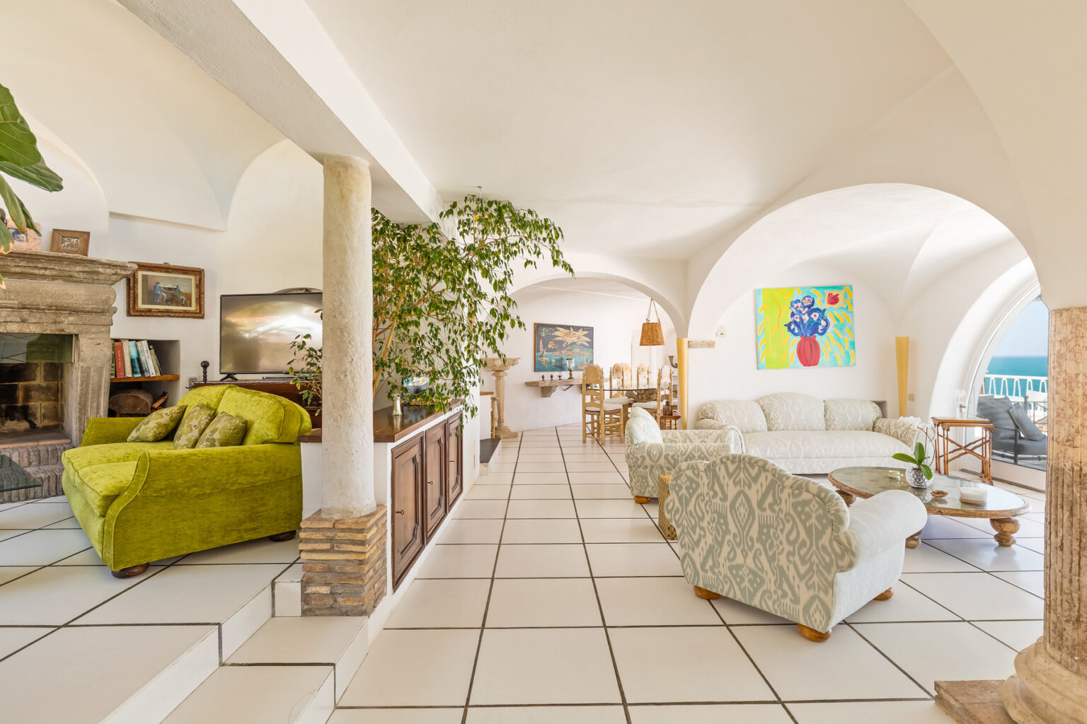 Bright and airy living room with modern decor, featuring a green armchair, white sofas, a stone fireplace, and large windows offering ocean views. The space includes plants and artwork, creating a welcoming atmosphere. | Only on SopranoVillas