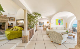 Bright and airy living room with modern decor, featuring a green armchair, white sofas, a stone fireplace, and large windows offering ocean views. The space includes plants and artwork, creating a welcoming atmosphere.