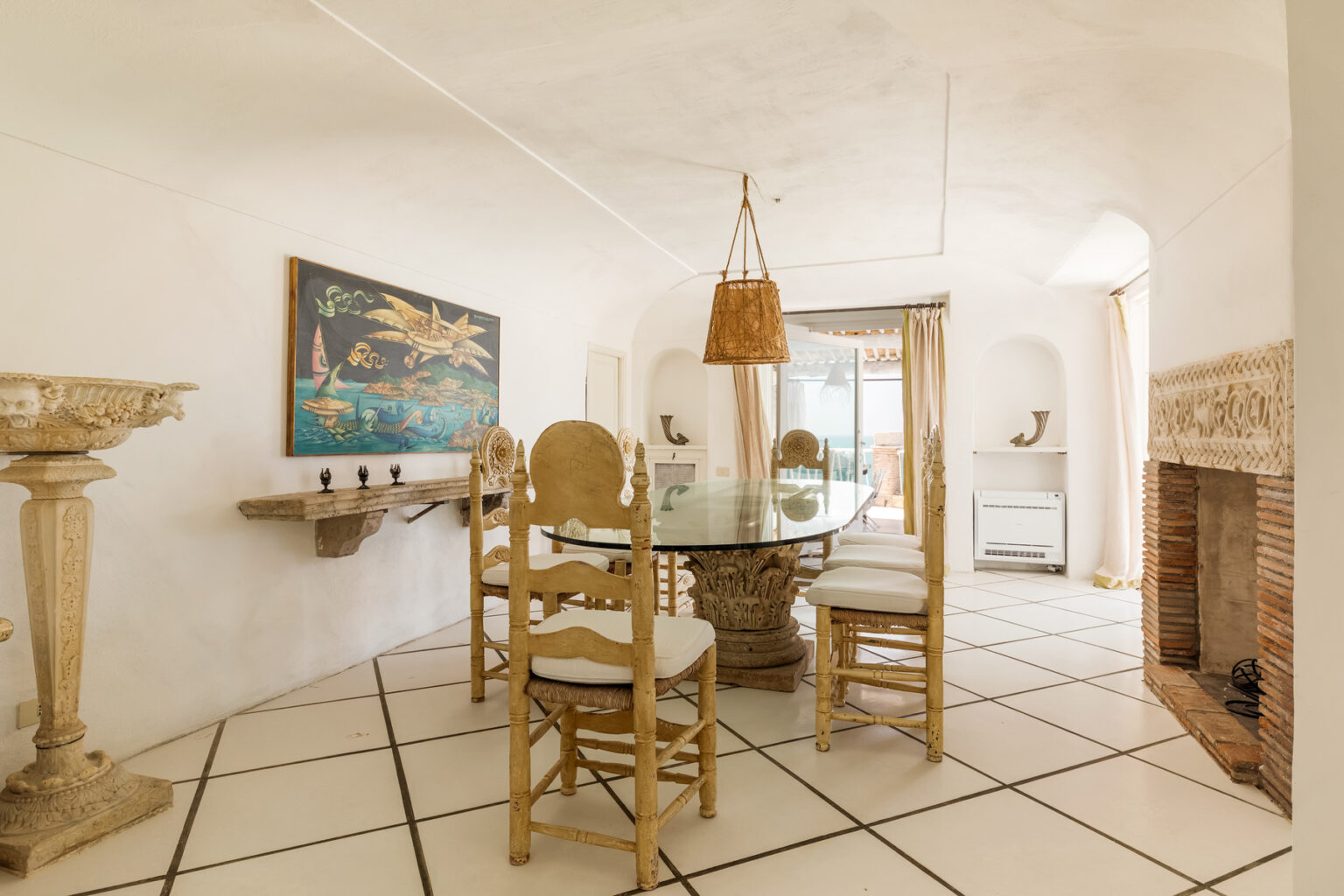 Bright and airy dining room featuring a glass table surrounded by elegant wooden chairs, a decorative wall painting, and a cozy fireplace, with large windows providing natural light and a view. | Only on SopranoVillas