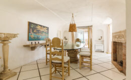 Bright and airy dining room featuring a glass table surrounded by elegant wooden chairs, a decorative wall painting, and a cozy fireplace, with large windows providing natural light and a view.