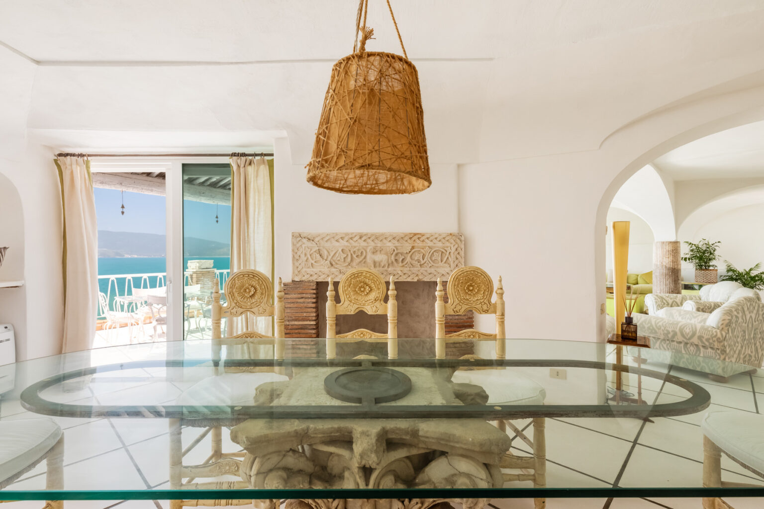 Bright and airy dining area featuring a glass table, rattan chairs, and a woven pendant light, with large windows showcasing a scenic view of the ocean. | Only on SopranoVillas