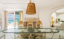 Bright and airy dining area featuring a glass table, rattan chairs, and a woven pendant light, with large windows showcasing a scenic view of the ocean.