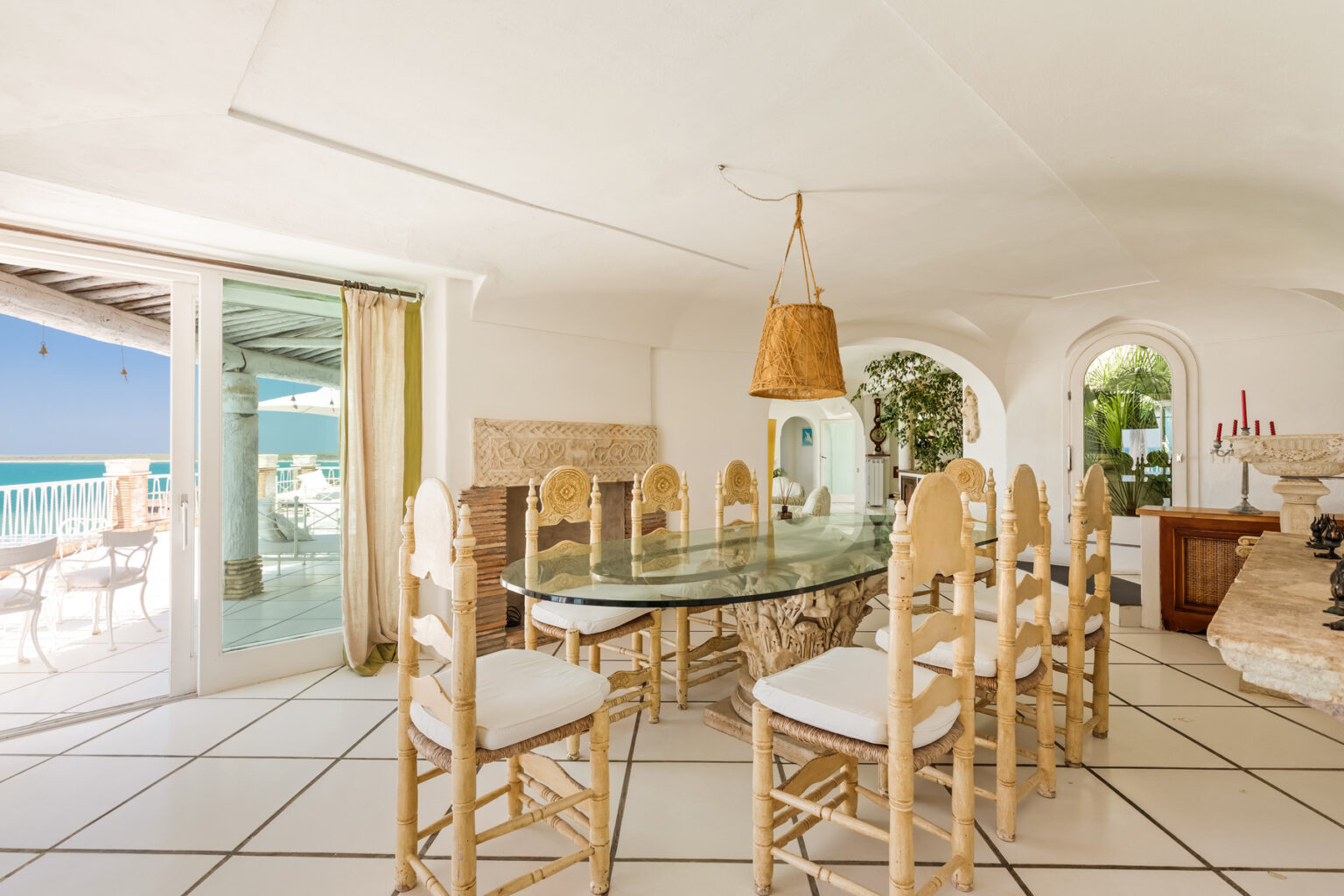 Bright and airy dining room featuring a glass table surrounded by light-colored chairs, with large windows offering a view of the ocean. The space is elegantly decorated with a warm pendant light and natural elements, creating a serene coastal atmosphere. | Only on SopranoVillas