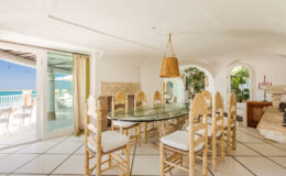 Bright and airy dining room featuring a glass table surrounded by light-colored chairs, with large windows offering a view of the ocean. The space is elegantly decorated with a warm pendant light and natural elements, creating a serene coastal atmosphere.
