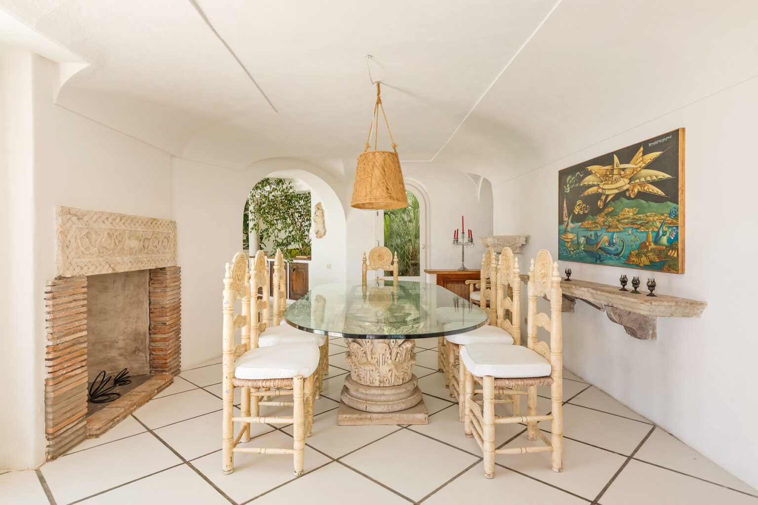Bright and airy dining room featuring a glass-top table surrounded by white wicker chairs, a rustic fireplace, and a colorful wall art piece, all set against a backdrop of white walls and tiled flooring. | Only on SopranoVillas