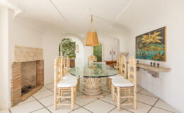 Bright and airy dining room featuring a glass-top table surrounded by white wicker chairs, a rustic fireplace, and a colorful wall art piece, all set against a backdrop of white walls and tiled flooring.
