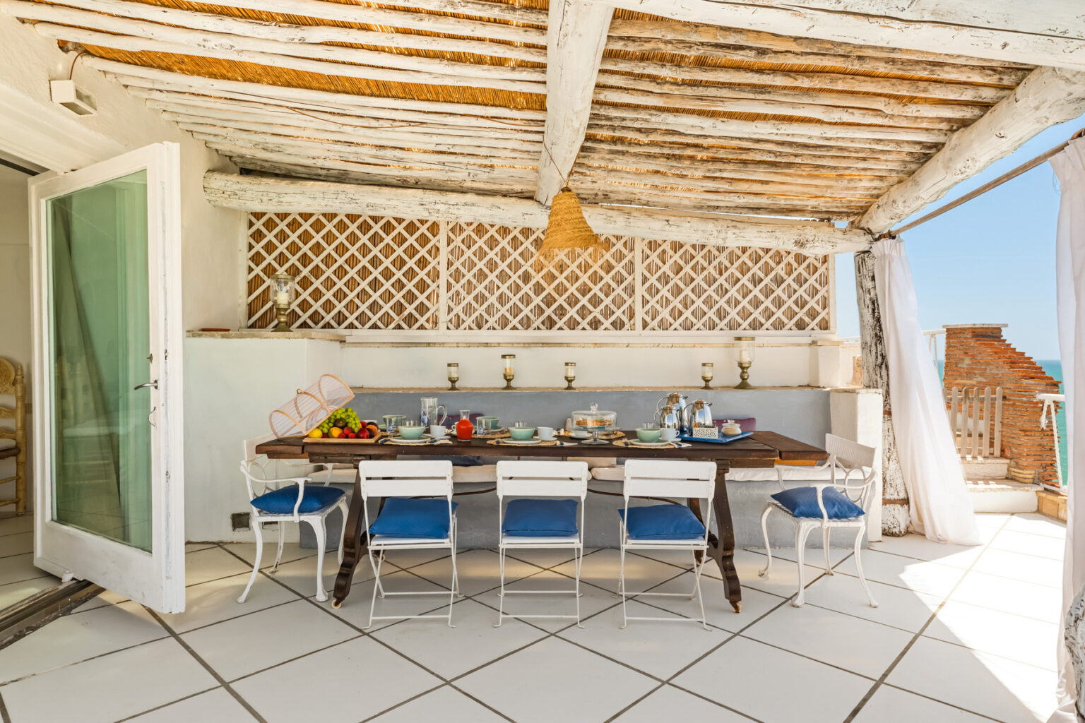 Stylish outdoor dining area featuring a large wooden table set with fresh fruit and tableware, surrounded by white chairs with blue cushions, under a rustic wooden ceiling, with a view of clear skies and a coastal landscape. Ideal for al fresco meals and gatherings. | Only on SopranoVillas