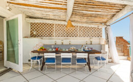 Stylish outdoor dining area featuring a large wooden table set with fresh fruit and tableware, surrounded by white chairs with blue cushions, under a rustic wooden ceiling, with a view of clear skies and a coastal landscape. Ideal for al fresco meals and gatherings.