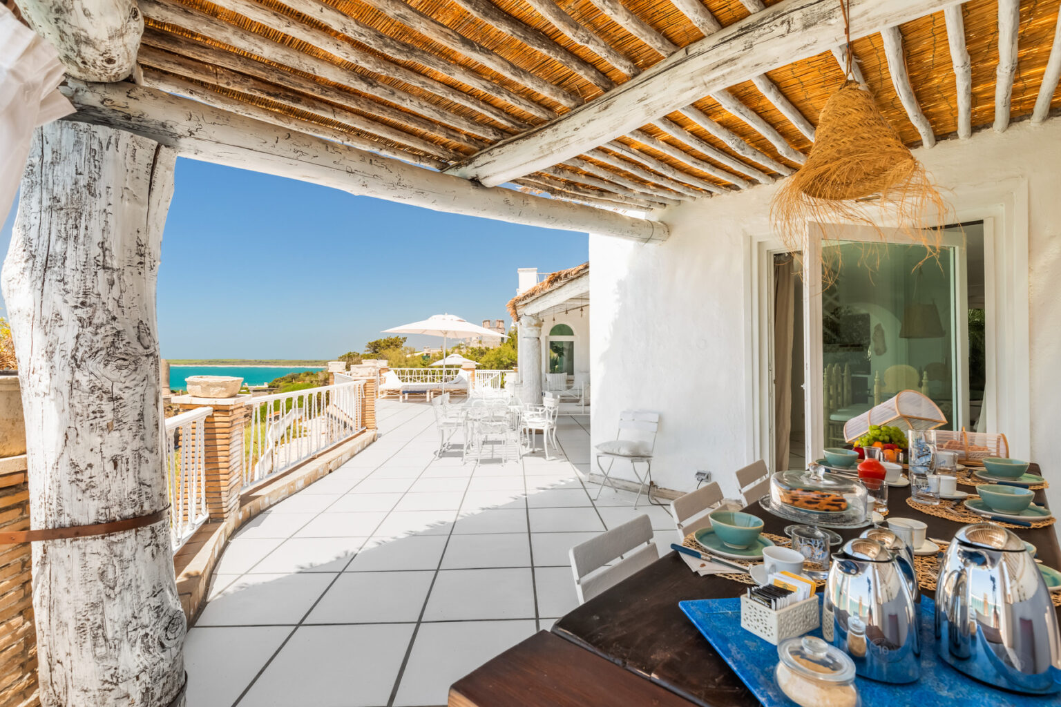 Outdoor dining area with a wooden pergola overlooking a seaside view, featuring a table set with elegant tableware and a relaxing atmosphere, ideal for enjoying meals in a coastal setting. | Only on SopranoVillas