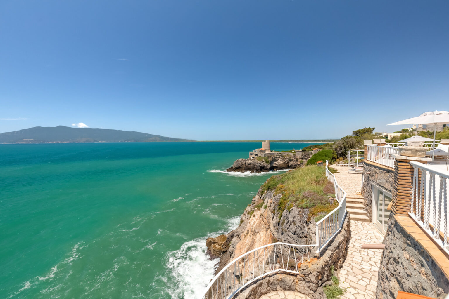 Scenic coastal view featuring turquoise waters, rocky cliffs, and a pathway along the shoreline, with a distant island visible under a clear blue sky. | Only on SopranoVillas