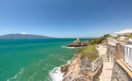 Scenic coastal view featuring turquoise waters, rocky cliffs, and a pathway along the shoreline, with a distant island visible under a clear blue sky.