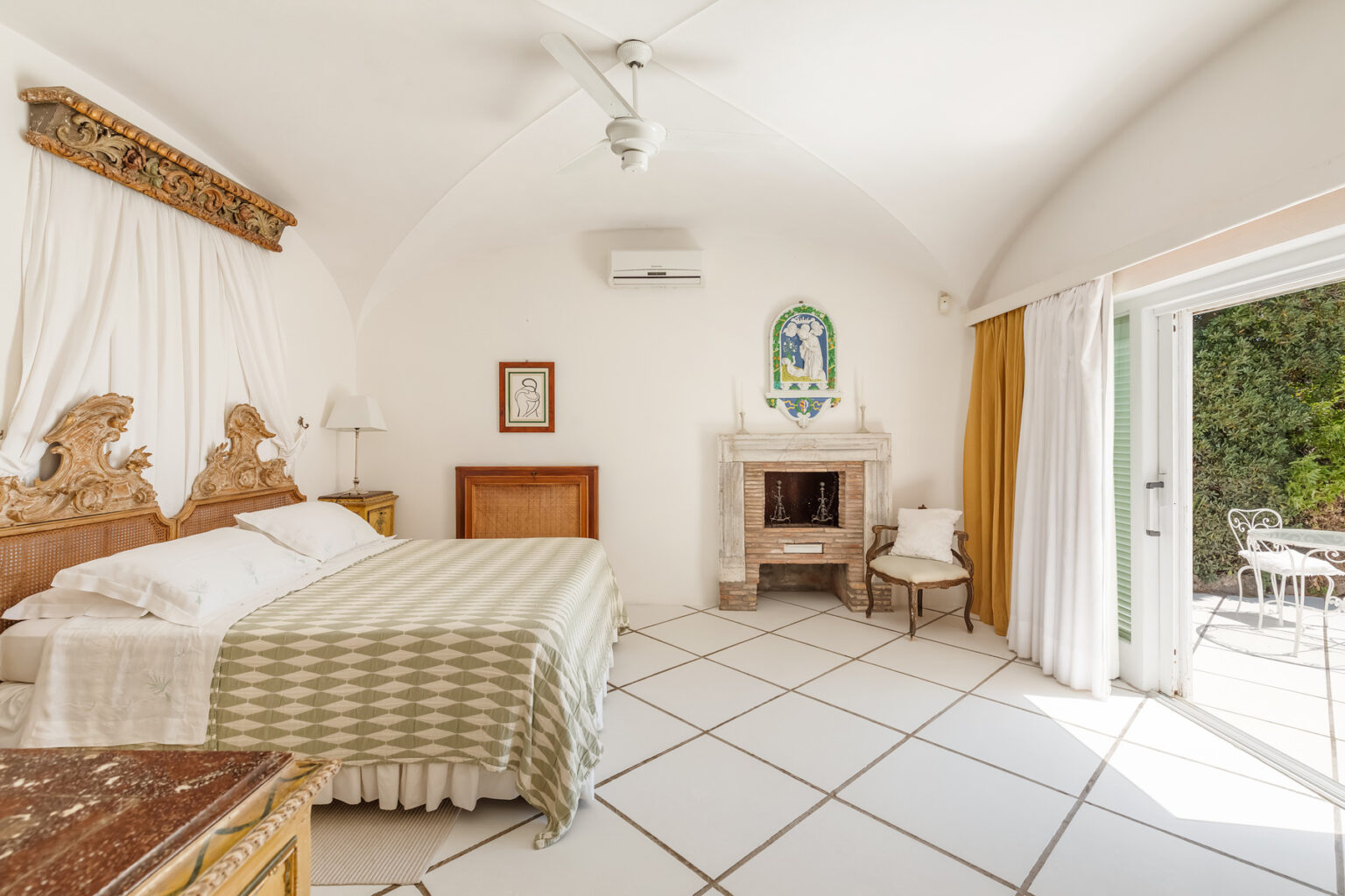 Cozy and elegant bedroom featuring a king-sized bed with a decorative headboard, white walls, and natural light streaming through sliding glass doors. The room includes a vintage fireplace, a small seating area, and a charming outdoor view, perfect for a relaxing retreat. | Only on SopranoVillas