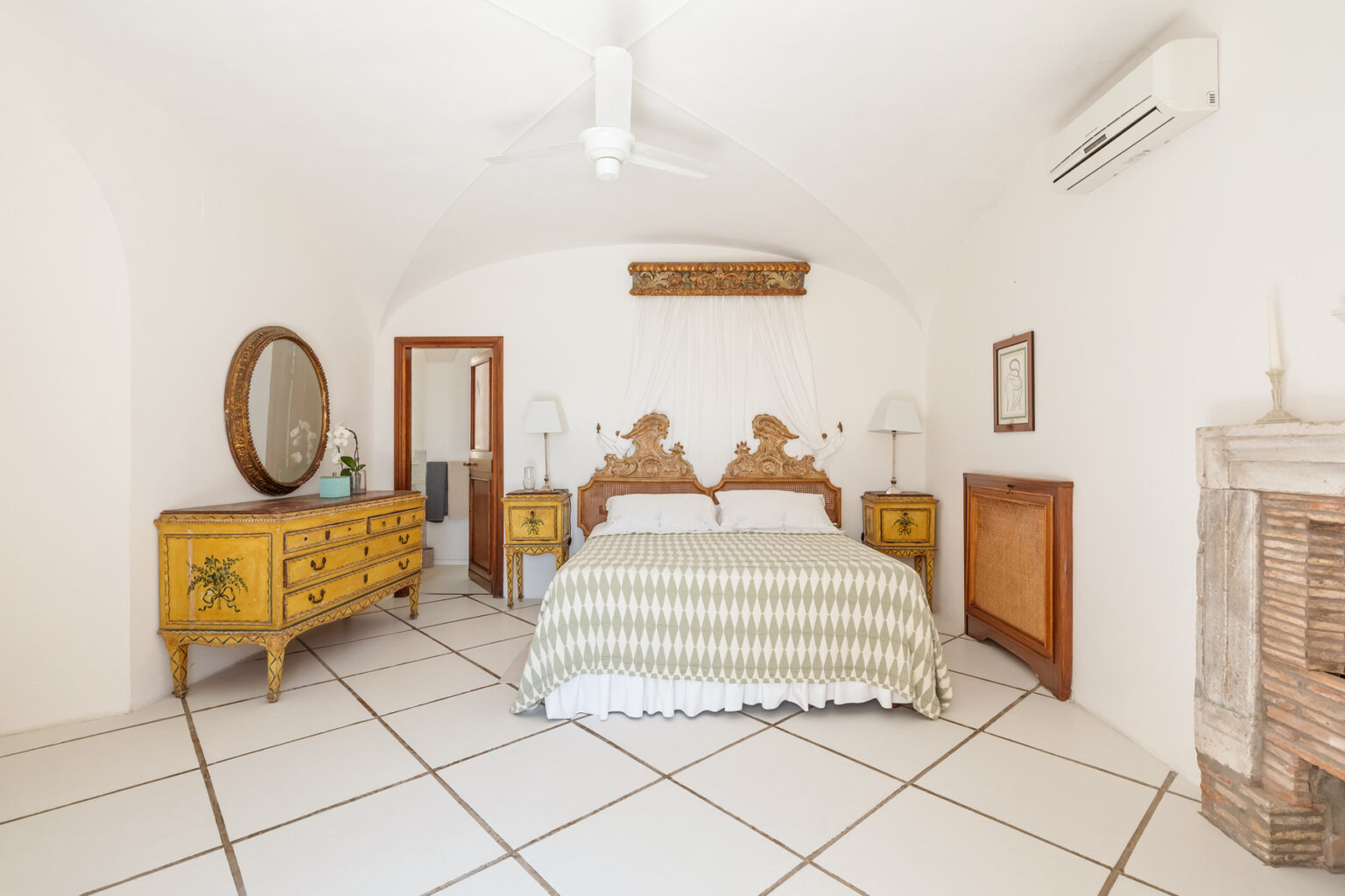 Bright and airy bedroom featuring a king-sized bed with a striped green and white comforter, vintage yellow wooden furniture, and a large round mirror. The room has white walls and tiled flooring, with natural light streaming in from a window. | Only on SopranoVillas