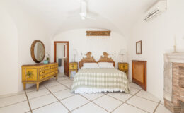 Bright and airy bedroom featuring a king-sized bed with a striped green and white comforter, vintage yellow wooden furniture, and a large round mirror. The room has white walls and tiled flooring, with natural light streaming in from a window.