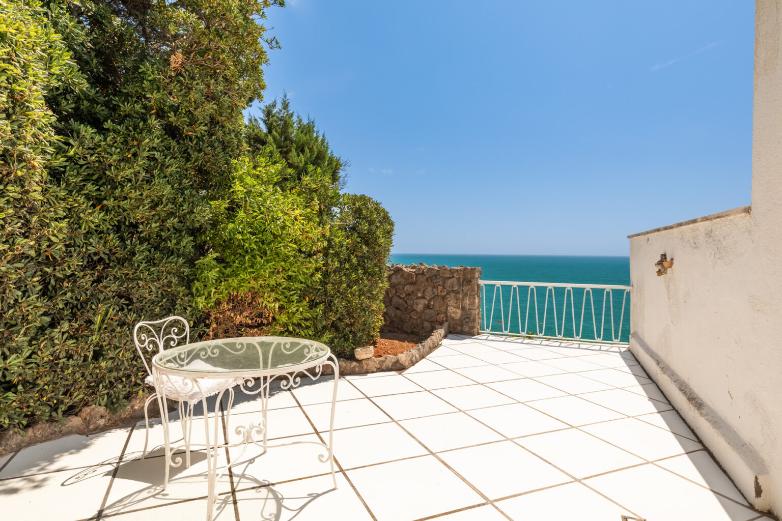 Cozy outdoor terrace with a white table and chairs overlooking the ocean, surrounded by lush greenery and a clear blue sky. Ideal for relaxation and enjoying scenic views. | Only on SopranoVillas