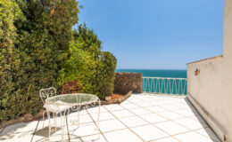 Cozy outdoor terrace with a white table and chairs overlooking the ocean, surrounded by lush greenery and a clear blue sky. Ideal for relaxation and enjoying scenic views.