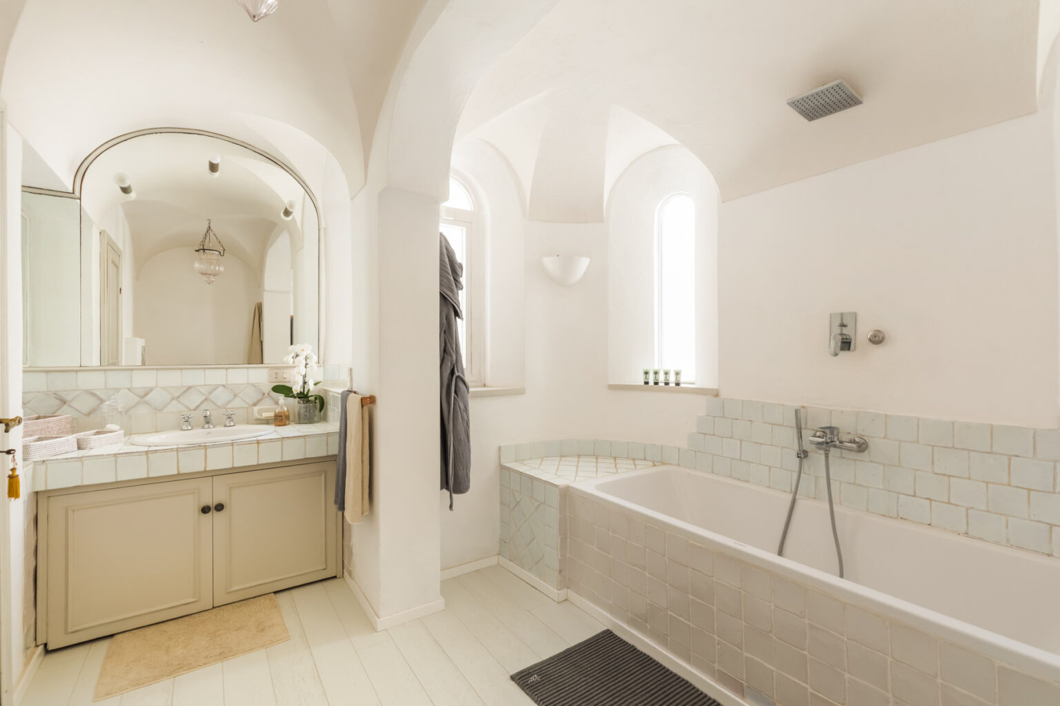 Modern bathroom design featuring a spacious layout with a freestanding bathtub, elegant light-colored tiles, and a double vanity with a large mirror. The room is illuminated by natural light from large windows, creating a bright and airy atmosphere. | Only on SopranoVillas