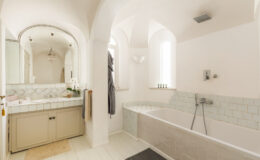 Modern bathroom design featuring a spacious layout with a freestanding bathtub, elegant light-colored tiles, and a double vanity with a large mirror. The room is illuminated by natural light from large windows, creating a bright and airy atmosphere.
