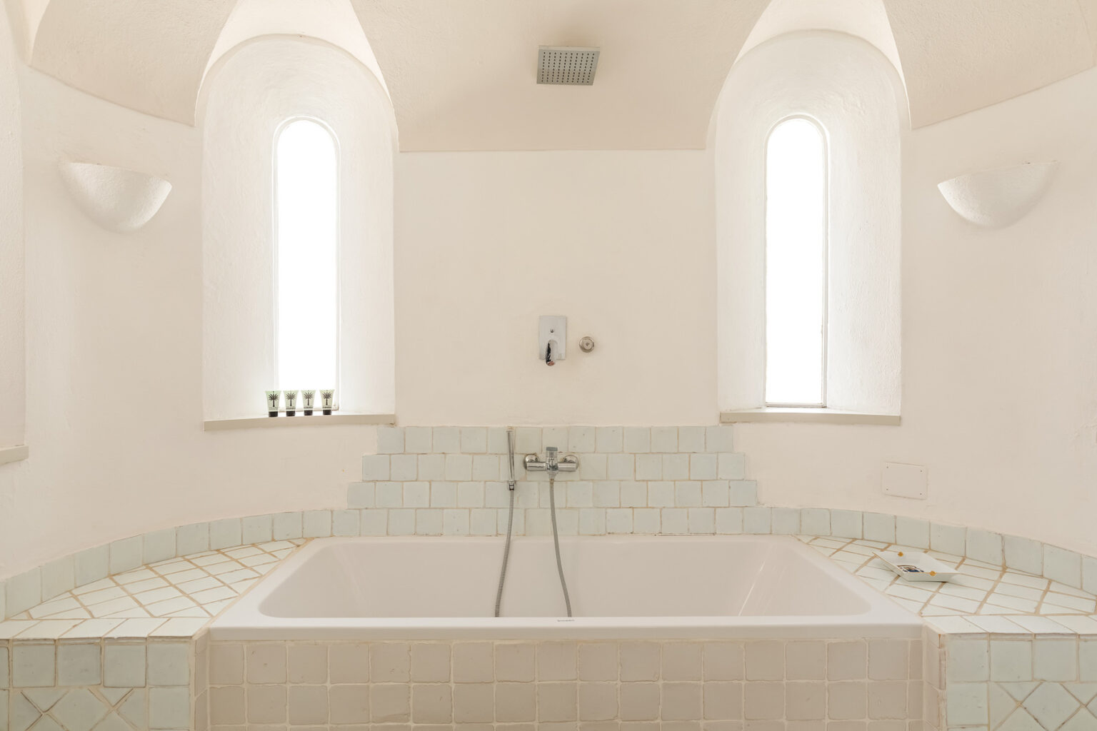 Modern bathroom interior featuring a spacious bathtub with a minimalist design, surrounded by light-colored tiles and elegant wall sconces. Natural light streams in through tall windows, creating a serene and inviting atmosphere. Ideal for relaxation and self-care. | Only on SopranoVillas