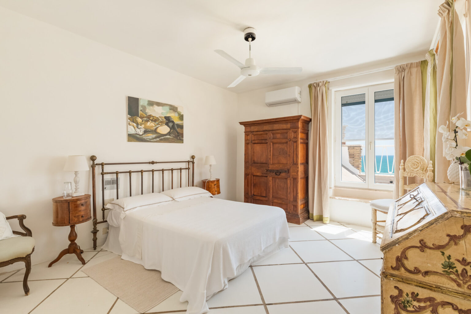 Cozy bedroom featuring a double bed with white linens, wooden furniture including a vintage wardrobe, and large windows that provide natural light and a view. The room is decorated in neutral tones with a ceiling fan for comfort. | Only on SopranoVillas