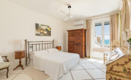 Cozy bedroom featuring a double bed with white linens, wooden furniture including a vintage wardrobe, and large windows that provide natural light and a view. The room is decorated in neutral tones with a ceiling fan for comfort.