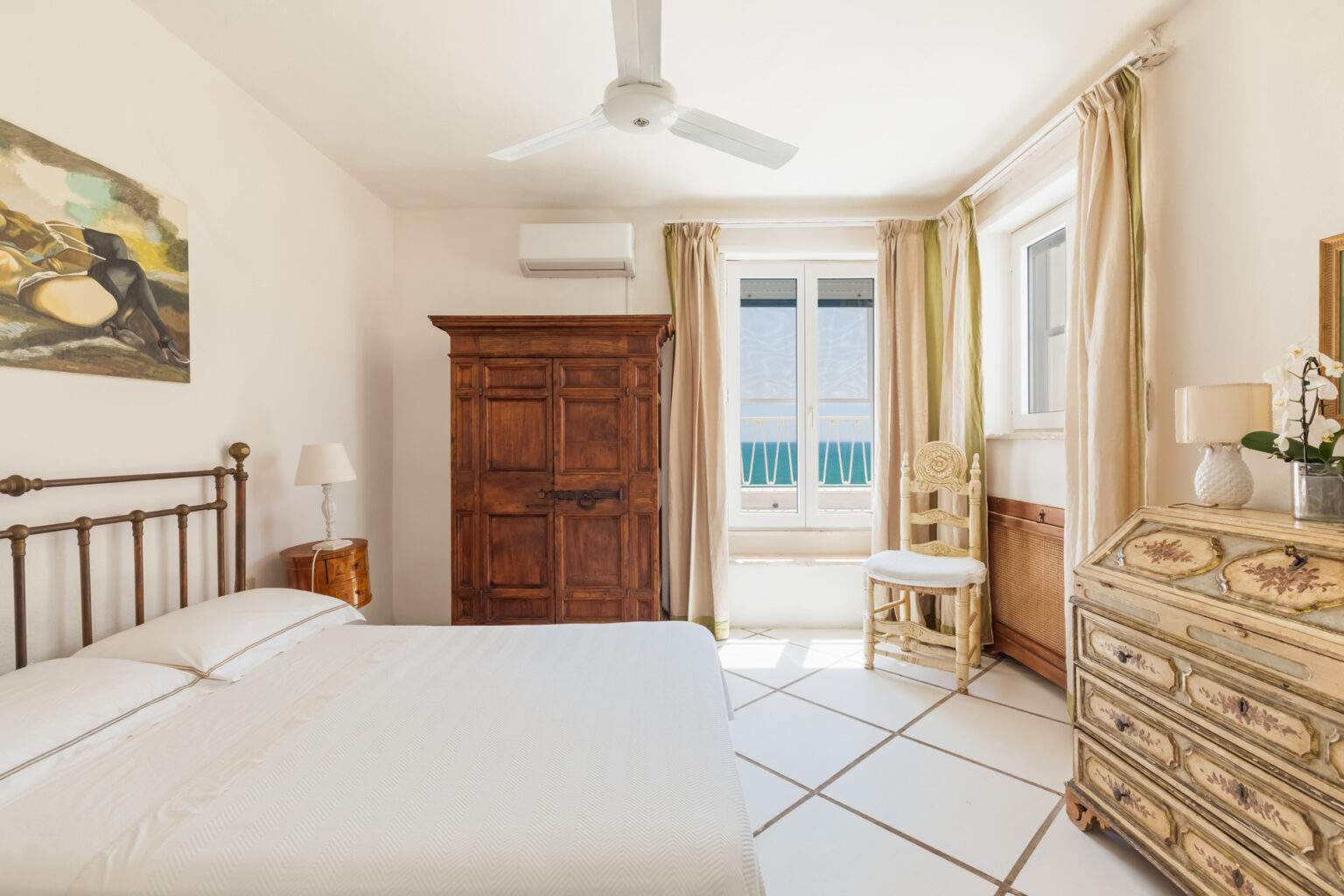 Bright and airy bedroom featuring a metal bed frame, vintage wooden wardrobe, decorative dresser, and large windows with scenic views. The room is accentuated by soft lighting and neutral tones, creating a cozy atmosphere. Ideal for relaxation and comfort. | Only on SopranoVillas