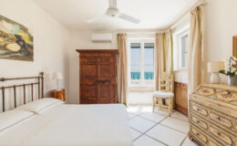 Bright and airy bedroom featuring a metal bed frame, vintage wooden wardrobe, decorative dresser, and large windows with scenic views. The room is accentuated by soft lighting and neutral tones, creating a cozy atmosphere. Ideal for relaxation and comfort.