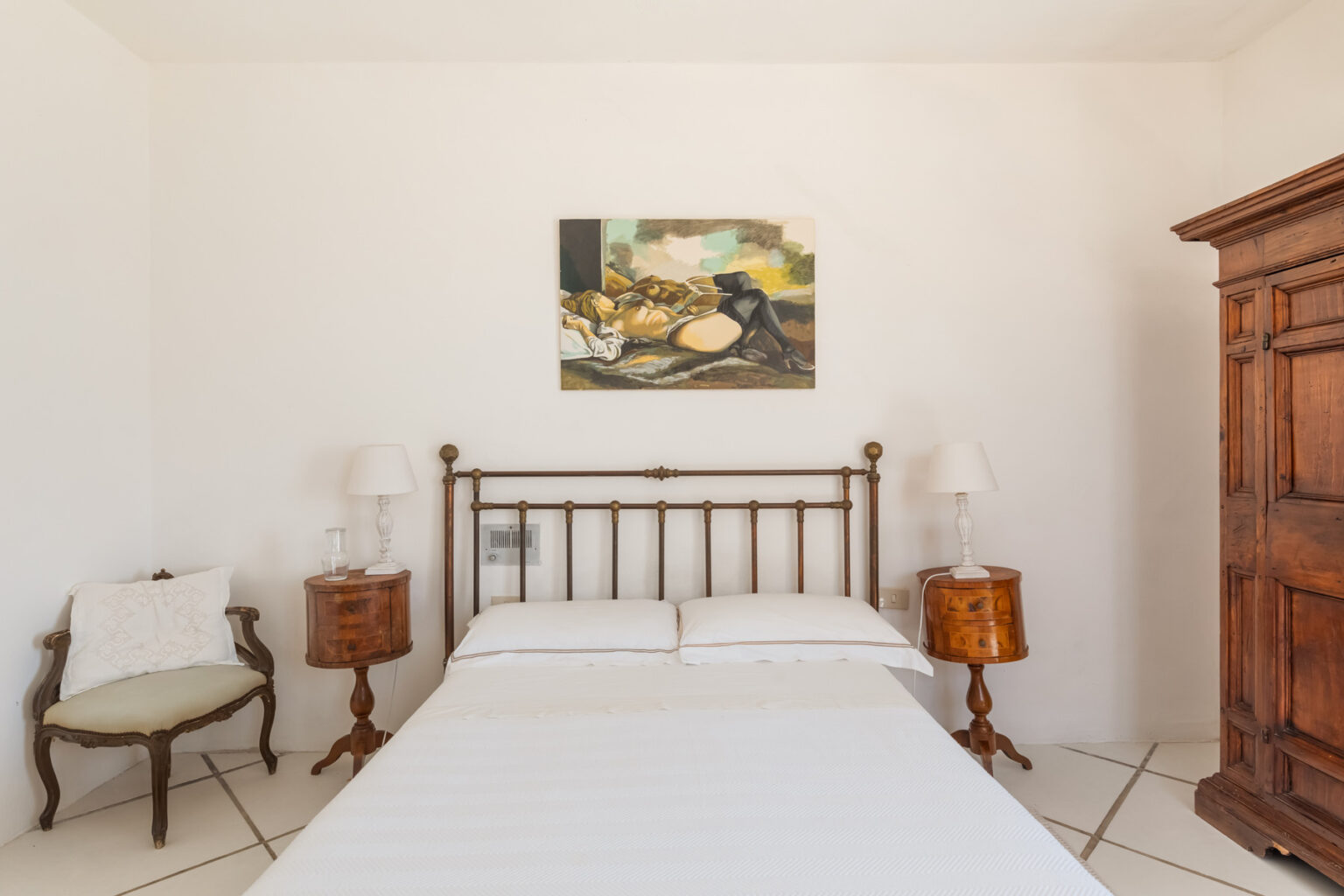 Cozy bedroom with a wooden bed frame, white bedding, vintage bedside tables, and an art piece on the wall, featuring a wooden wardrobe and a small chair in a light-filled space. | Only on SopranoVillas