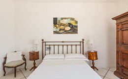 Cozy bedroom with a wooden bed frame, white bedding, vintage bedside tables, and an art piece on the wall, featuring a wooden wardrobe and a small chair in a light-filled space.