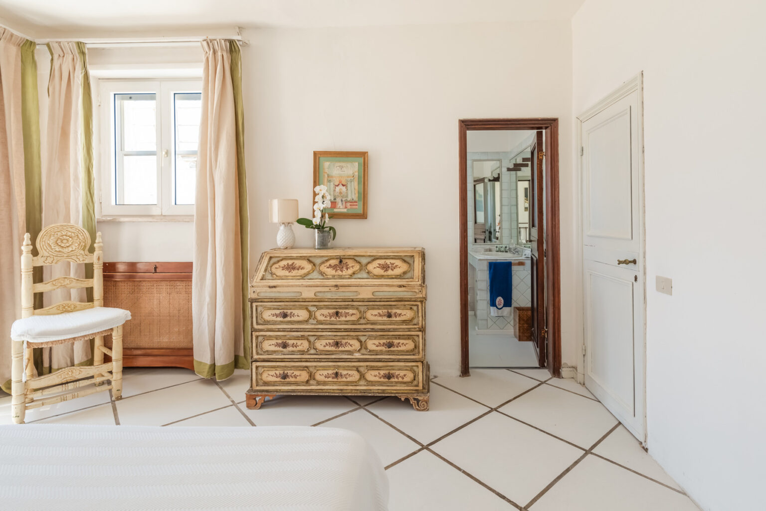 Bright and airy bedroom featuring a vintage wooden dresser, a white chair, and large windows with light curtains. A mirror reflects the adjoining bathroom, enhancing the room's spacious feel. | Only on SopranoVillas