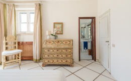 Bright and airy bedroom featuring a vintage wooden dresser, a white chair, and large windows with light curtains. A mirror reflects the adjoining bathroom, enhancing the room's spacious feel.