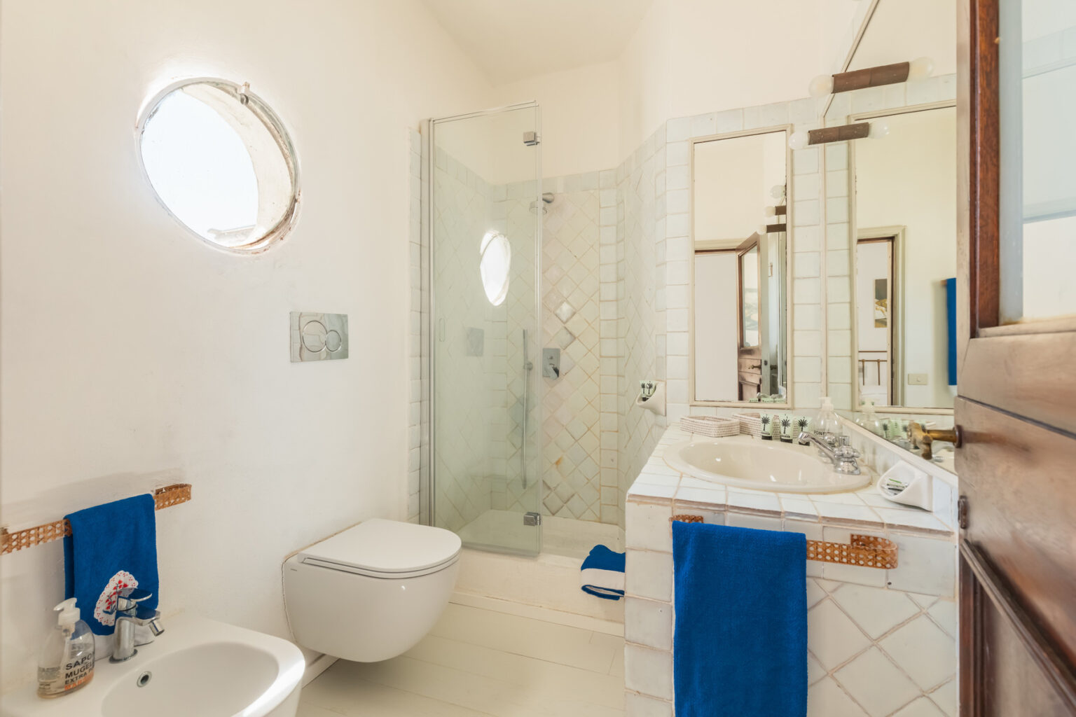 Modern bathroom featuring a glass shower, white tiled walls, and a round window, with a toilet and a stylish sink area adorned with blue towels. | Only on SopranoVillas