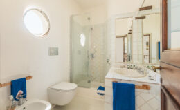 Modern bathroom featuring a glass shower, white tiled walls, and a round window, with a toilet and a stylish sink area adorned with blue towels.