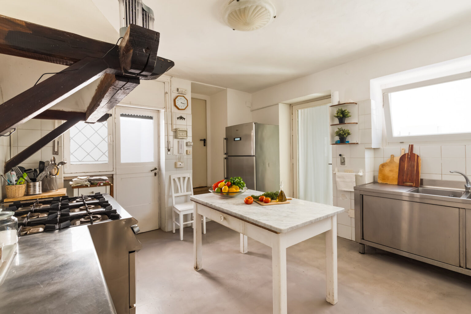 Bright and modern kitchen featuring a marble-topped table with fresh vegetables, stainless steel appliances, and wooden accents. The space includes a gas stove, a large refrigerator, and ample natural light from windows, creating an inviting atmosphere for cooking and dining. | Only on SopranoVillas